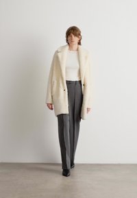 Kreemivärvi faux fur jakk, millel on mugav lõige, notšitud krae ja kaks esist pocketit, kombineerituna hallide kohandatud pükstega ja mustade kingadega.