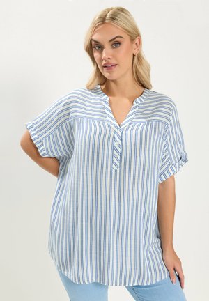 Mujer rubia con blusa blanca holgada de manga corta con rayas azules verticales y horizontales, combinada con jeans azul claro, de pie con la mano en la cadera.