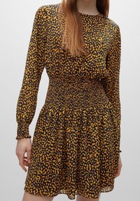 Zwarte jurk met een gele dierenprint, lange mouwen en een smock taille. De stof is lichtgewicht met een vloeiende silhouet.