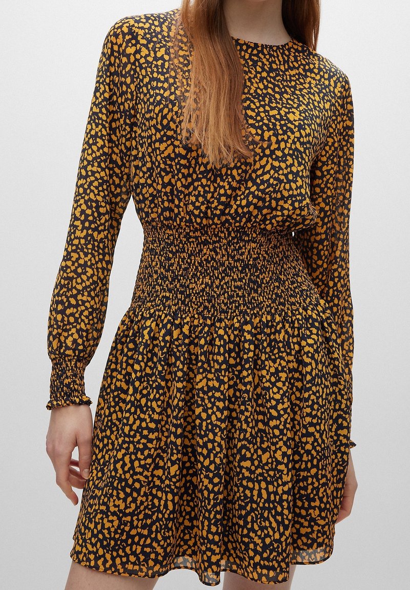 Zwarte jurk met een gele dierenprint, lange mouwen en een smock taille. De stof is lichtgewicht met een vloeiende silhouet.