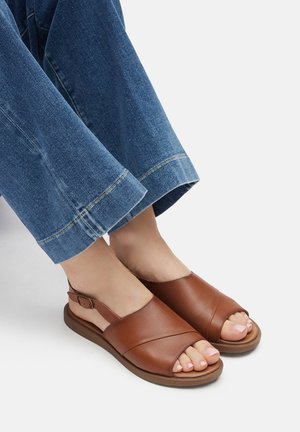 Hnědé kožené sandály s otevřenou špičkou a designem křížových pásků. Nízký zaoblený podpatek s nastavitelným páskem kolem kotníku. Nošeno s širokými džínovými kalhotami.