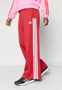 Piros sportnadrág három fehér csíkkal az oldalán és egy Adidas logóval. Puha anyag, laza fazon, fekete sportcipőkkel párosítva.