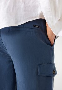 Pantalons cargo navy en tissu léger, dotés d'une poche latérale avec un bouton et d'une étiquette logo noire "GUESS" visible.