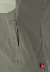 Šedá bunda se světlým šedým panelem, texturovanou tkaninou, boční kapsou a červenou značkovou etiketou poblíž lemu. Jednoduchý, funkční design.