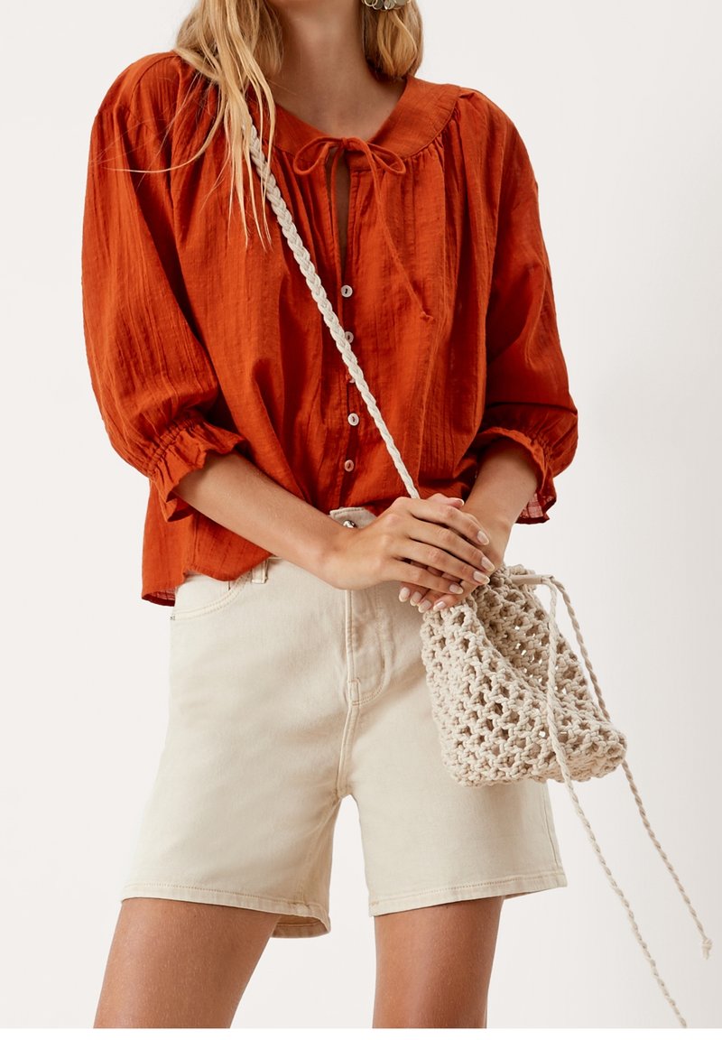 Femme portant un chemisier boutonné couleur rouille avec un nœud papillon, un short crème et tenant un sac bandoulière tricoté crème.