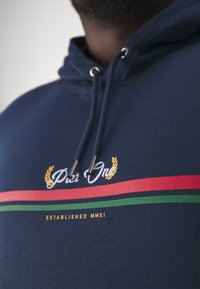 Sudadera azul marino con cordón y detalles de metal. En la parte frontal tiene texto bordado y franjas horizontales en rojo y verde.