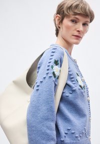 Ανοιχτό μπλε πλεκτό πουλόβερ με κεντήματα λουλουδιών, υφή επιφάνειας και πέρλες κουμπιά. Μια κρεμ τσάντα ώμου είναι περασμένη στον βραχίονα.