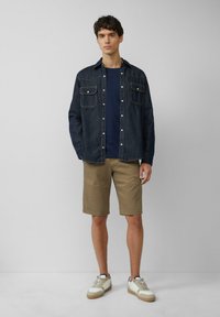Dunkelblaues Denim-Hemd mit zwei Taschen auf der Vorderseite, getragen über einem marineblauen T-Shirt und kombiniert mit hellbraunen Shorts und weißen Sneakers.