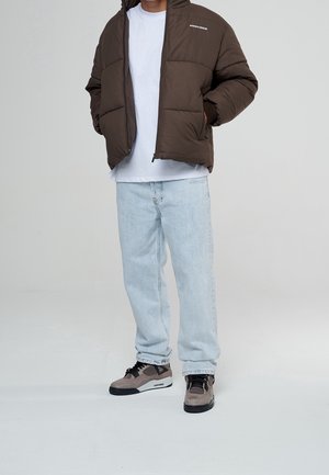 Brun oppustelig jakke med hætte, hvid t-shirt under, lyseblå jeans og grå sneakers med sorte detaljer, stående på en hvid baggrund.