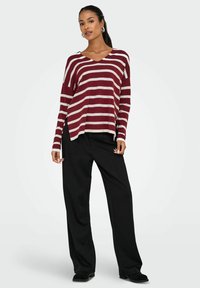 Pull rayé bordeaux et crème avec un col en V, des manches longues surdimensionnées et des fentes sur les côtés, associé à un pantalon noir ample et des chaussures noires.