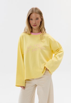 Jonge vrouw met lang blond haar, gekleed in een gele trui met roze halslijn en de tekst "wees echt, niet perfect", gecombineerd met lichtbeige broek.