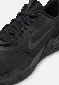 Zapatilla deportiva negra con tejido texturizado, zonas reforzadas y un logo de Nike en gris. Presenta un cuello acolchado y cordones planos para un ajuste seguro.