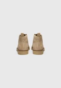 Botines de ante beige con un tacón bajo, que presentan una puntera redondeada, un tirador en la parte superior y costuras contrastantes a lo largo de las costuras.