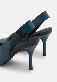 Escarpins slingback en cuir bleu avec bride élastique noire et détail logo sur fond blanc.
