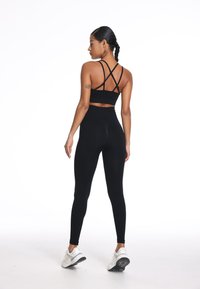 Zwart sport-bh met gekruiste bandjes, gecombineerd met hoge zwarte leggings. Model draagt witte sportschoenen, staande op een effen achtergrond.