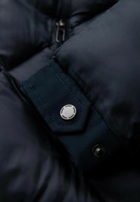 Marinblå puffjacka med en slät textur, utrustad med hexagonalt tygdetalj och en silverfärgad tryckknapp med ingraverat varumärkesnamn.