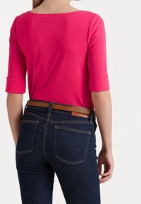 Tajmad rosa topp med trekvartslånga ärmar, kombinerad med mörkblå jeans som har ett brunt bälte och stickningsdetaljer på fickorna.