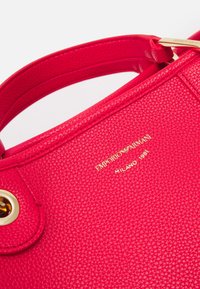 Czerwona torebka ze skóry o fakturze, z złotym emblematem z napisem „Emporio Armani” i „Milano 1981”, posiadająca zaokrąglony górny kształt i solidny uchwyt.