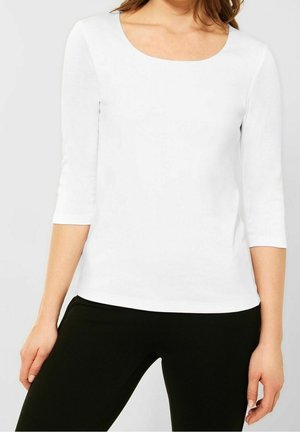 Femme portant un haut blanc uni à manches 3/4 et un pantalon noir, debout devant un fond neutre.