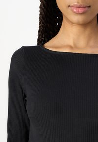 Top noir à manches longues en côtes, avec un décolleté large et rond, présentant une texture lisse et dévoilant le teint de peau au niveau du col.