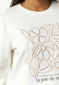 Vit sweatshirt med beige floralt broderi och texten "la joie de vivre" nära bröstet, bärs av en person, beskuren under hakan.