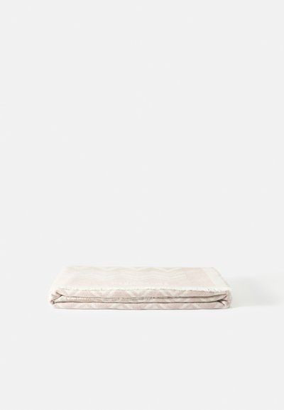 Couverture beige pliée avec des motifs subtils et des bords à franges, posée à plat sur une surface blanche.
