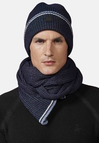 Berretto di lana blu navy con striscia bianca; sciarpa di lana abbinata drappeggiata attorno al collo, tessuto testurizzato, indossata sopra una maglietta nera.