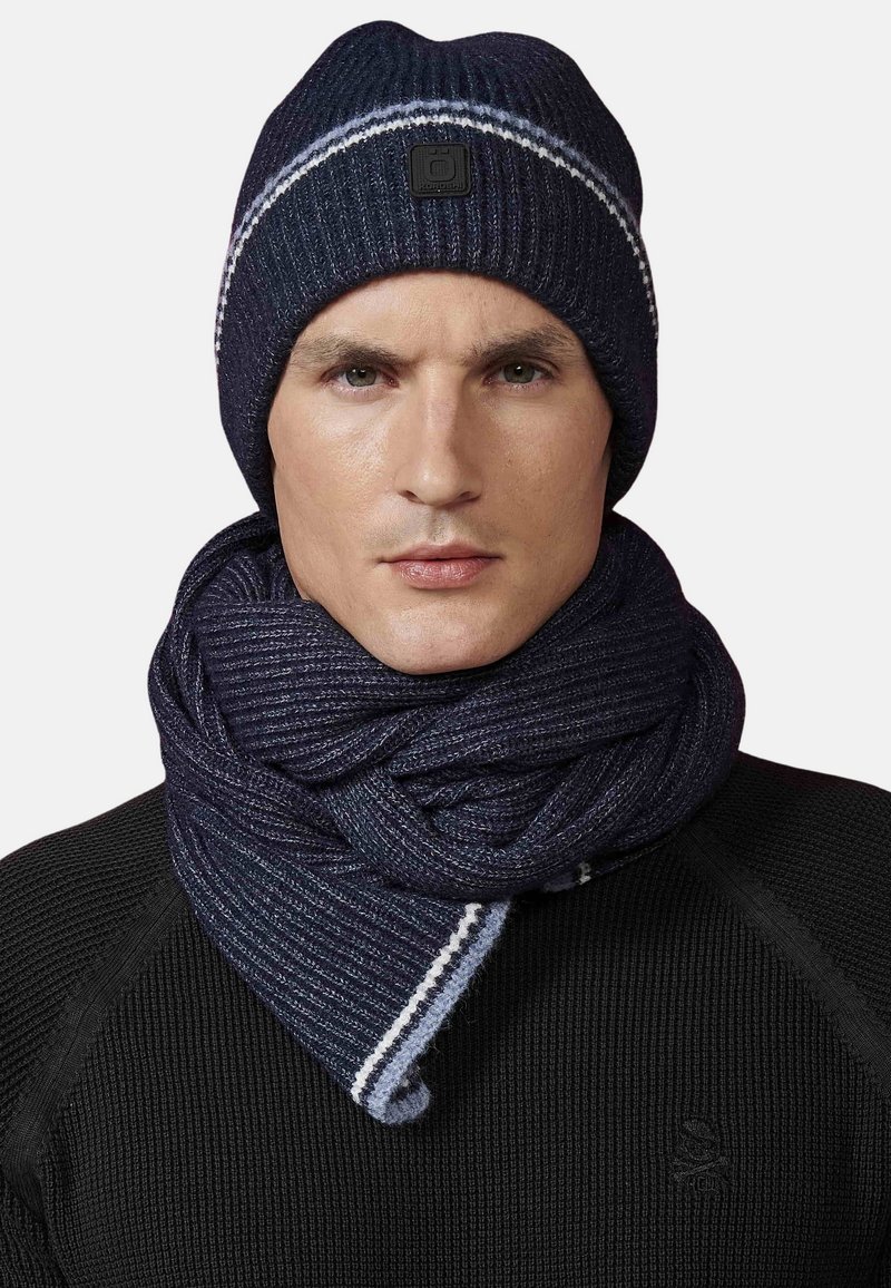 Berretto di lana blu navy con striscia bianca; sciarpa di lana abbinata drappeggiata attorno al collo, tessuto testurizzato, indossata sopra una maglietta nera.