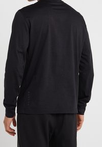 Långärmad svart t-shirt i bomull, med rund halsringning och en logotyp tryckt vertikalt på vänster sida nära fållen.