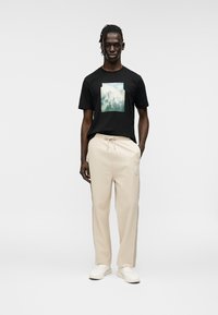 Μαύρο T-shirt με πράσινο και μπλε γραφικό σχέδιο, συνδυασμένο με μπεζ φόρμα. Το σύνολο έχει άνετη εφαρμογή και λευκά sneakers.