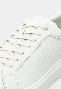 White leather sneaker with textured surface featuring a repeated logo pattern, flat laces, and a smooth rubber sole.