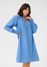 Robe bleue à manches longues, ample, en tissu texturé, avec un décolleté en V et une fermeture à nouer, ainsi que des poignets froncés.