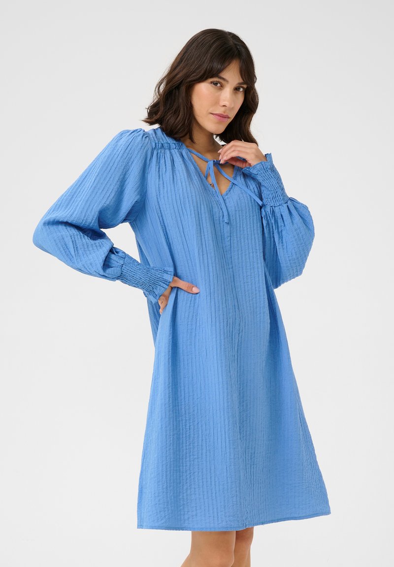 Robe bleue à manches longues, ample, en tissu texturé, avec un décolleté en V et une fermeture à nouer, ainsi que des poignets froncés.