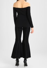 Schwarzes Off-Shoulder-Top und schwarze, ausgestellte Jeans mit Akzentnähten; taillierte Passform mit glatter Textur; High Heels.