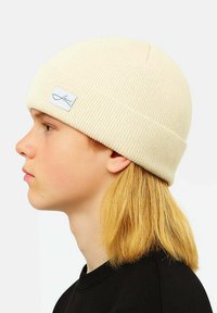 Cream-colored knitted beanie with a ribbed texture and a folded cuff. Features a small, rectangular label with cursive text.
