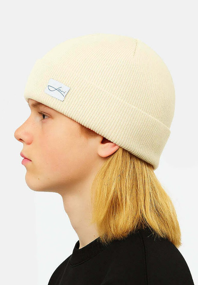Cream-colored knitted beanie with a ribbed texture and a folded cuff. Features a small, rectangular label with cursive text.