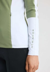 Lättviktig atletisk topp med en grön och vit färgblockdesign, slät textur, dragkedjedetalj och logga på ärmen.