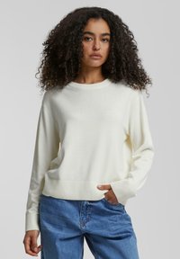 Pull en tricot de couleur crème avec un col rond, des poignets et un ourlet côtelés, et des manches longues, associé à un jean en denim bleu.