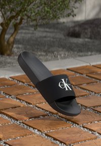 Black slide sandal with a wide strap displaying a white "Calvin Klein" logo. Smooth texture, contoured footbed, raised edges, rubber sole.