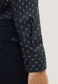 Camicia a maniche lunghe in tessuto blu scuro con una piccola stampa paisley bianca e azzurra. Presenta un polsino con bottoni e un colletto a contrasto.