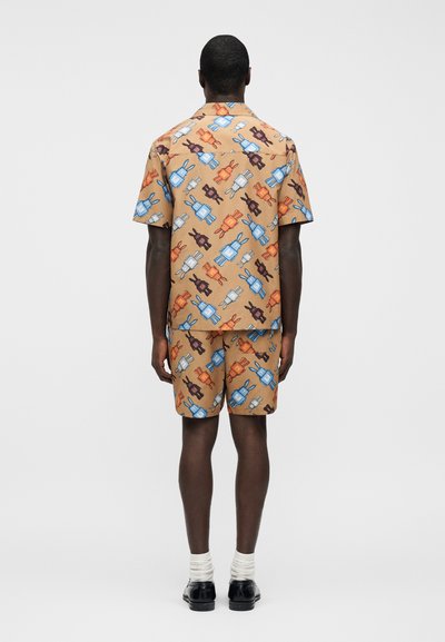 Homme portant une chemise à manches courtes et un short avec un fond beige et un motif coloré de robot et de lapin, debout de dos à la caméra.