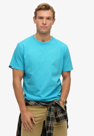Light blue cotton t-shirt with short sleeves and a crew neckline, featuring a small logo on the chest. Paired with khaki pants and a plaid shirt tied at the waist.