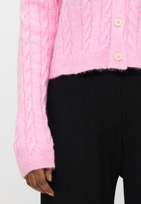 Ροζ πλεκτή ζακέτα με κοντό σχέδιο, μακριά μανίκια και δύο κουμπιά. Μαύροι ριμπ υφασμένοι παντελόνες είναι μερικώς ορατοί από κάτω.