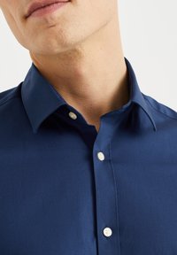 Dolní část obličeje a krku muže v tmavě modré košili s bílými knoflíky, límec rozepnutý, na jednoduchém bílém pozadí.