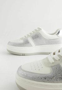 Weiße Sneakers mit silbernen Mesh-Akzenten, gepolstertem Kragen und flachen Schnürsenkeln. Verfügt über eine dicke Plattformsohle mit durchbrochenen Zehen Details.