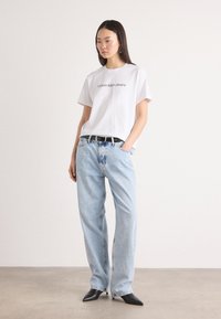 Balta medvilninė T-marškinė su "Calvin Klein Jeans" logo, derinama su šviesiai mėlynomis aukštu juosmeniu džinsais ir juodomis smailėmis aukštakulnėmis.