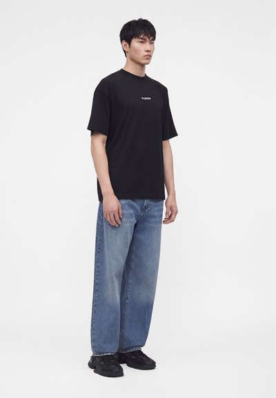 Νέος άνδρας που φοράει μαύρο T-shirt Balenciaga, μπλε φαρδιά τζιν και μαύρα αθλητικά παπούτσια, μπροστά σε απλό λευκό φόντο.