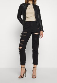 Vrouw in gescheurde zwarte jeans, beige gebreide top, zwart leren jack en zwarte hakken, staand tegen een witte achtergrond.