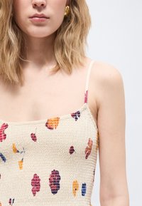 Νεαρή γυναίκα με ξανθά μαλλιά φοράει ένα φόρεμα σε εκρού χρώμα με λάστιχο στη μέση, διακοσμημένο με μοτίβα φύλλων σε πορτοκαλί, κόκκινο και μπλε, και ένα χρυσό σκουλαρίκι.