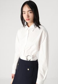Camicia bianca con bottoni e maniche lunghe, abbinata a una gonna blu navy con due tasche frontali e un dettaglio a cintura circolare.